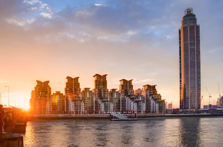 The image depicts a cityscape at sunset, featuring a row of modern residential buildings with distinctive stepped terraces and large glass windows, situated along the riverbank. A tall, cylindrical sk