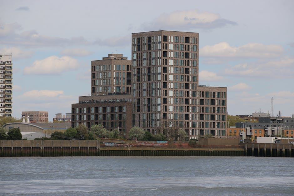 The image depicts a cityscape at sunset, featuring a row of modern residential buildings with distinctive stepped terraces and large glass windows, situated along the riverbank. A tall, cylindrical sk