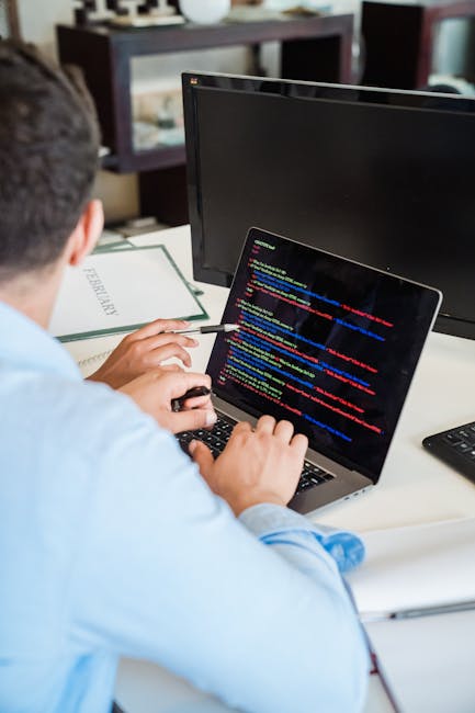 A person working on a MacBook laptop with a black screen displaying lines of colorful code, seated at a dark table in a well-lit indoor setting. The individual's hands are typing on the keyboard, and 