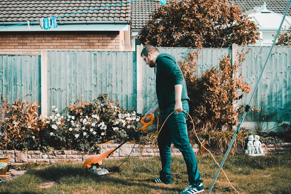 A man wearing glasses, a dark hoodie, and jeans is operating an orange electric lawn trimmer in a well-maintained backyard garden. He is trimming the grass along a flower bed that contains white and b